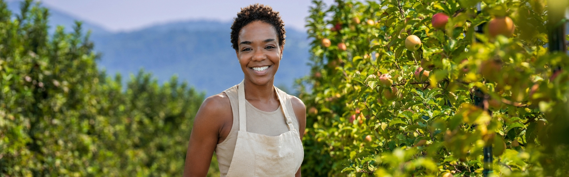 Stephanie Willis standing in an orchard, smiling, apple trees on either side